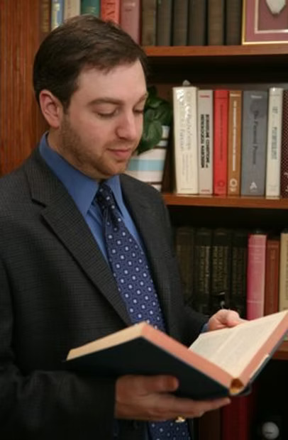 Dr. Aaron Goldner, in a jacket and tie, reading a book in front of a shelf of psychology texts